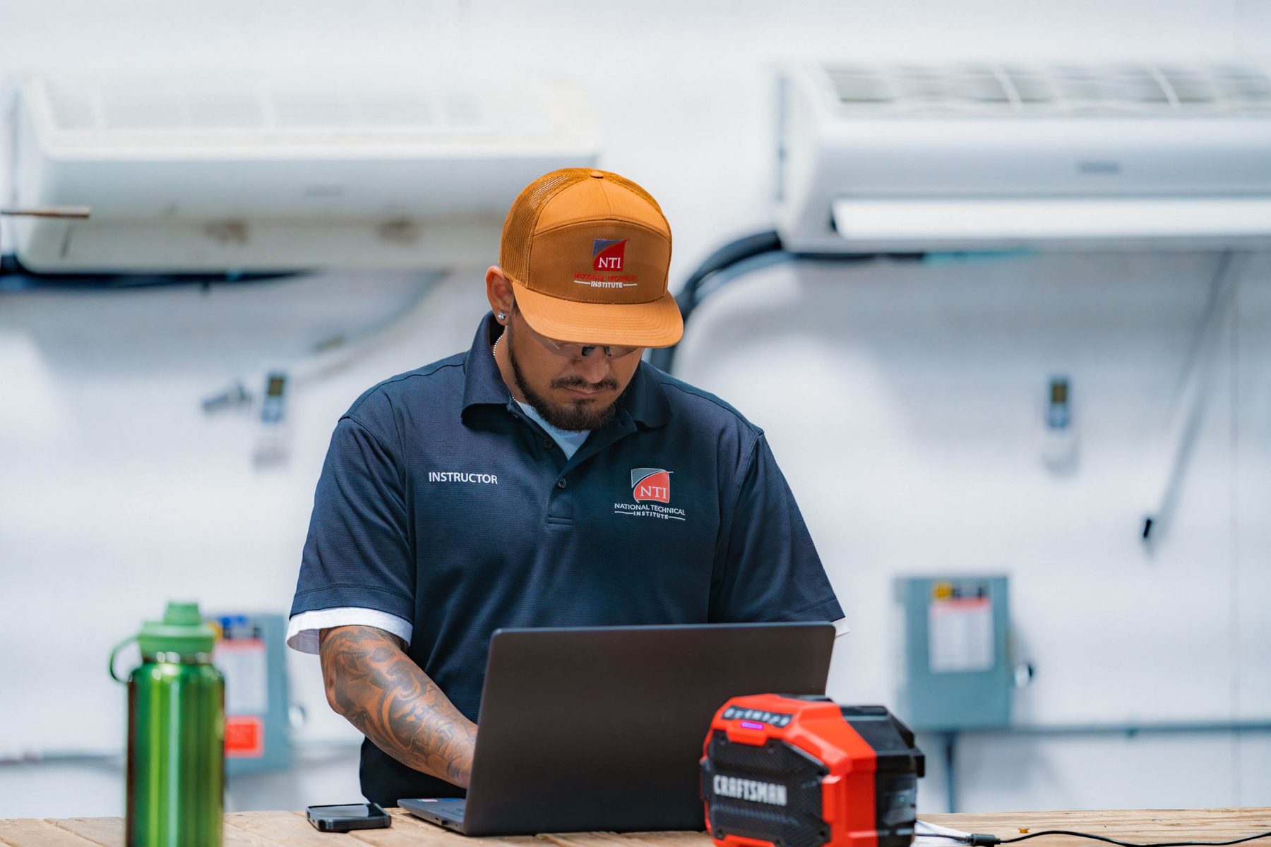 An NTI instructor at a laptop in front of a wall of HVAC training units