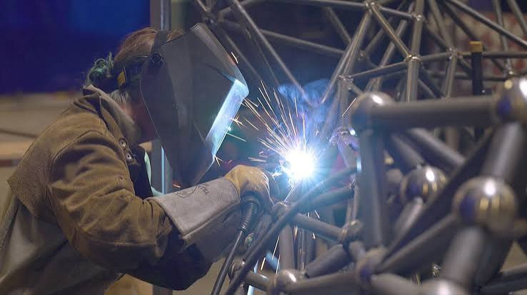 A welder in a leather jacket works on a large steel sculpture frame, sparks flying
