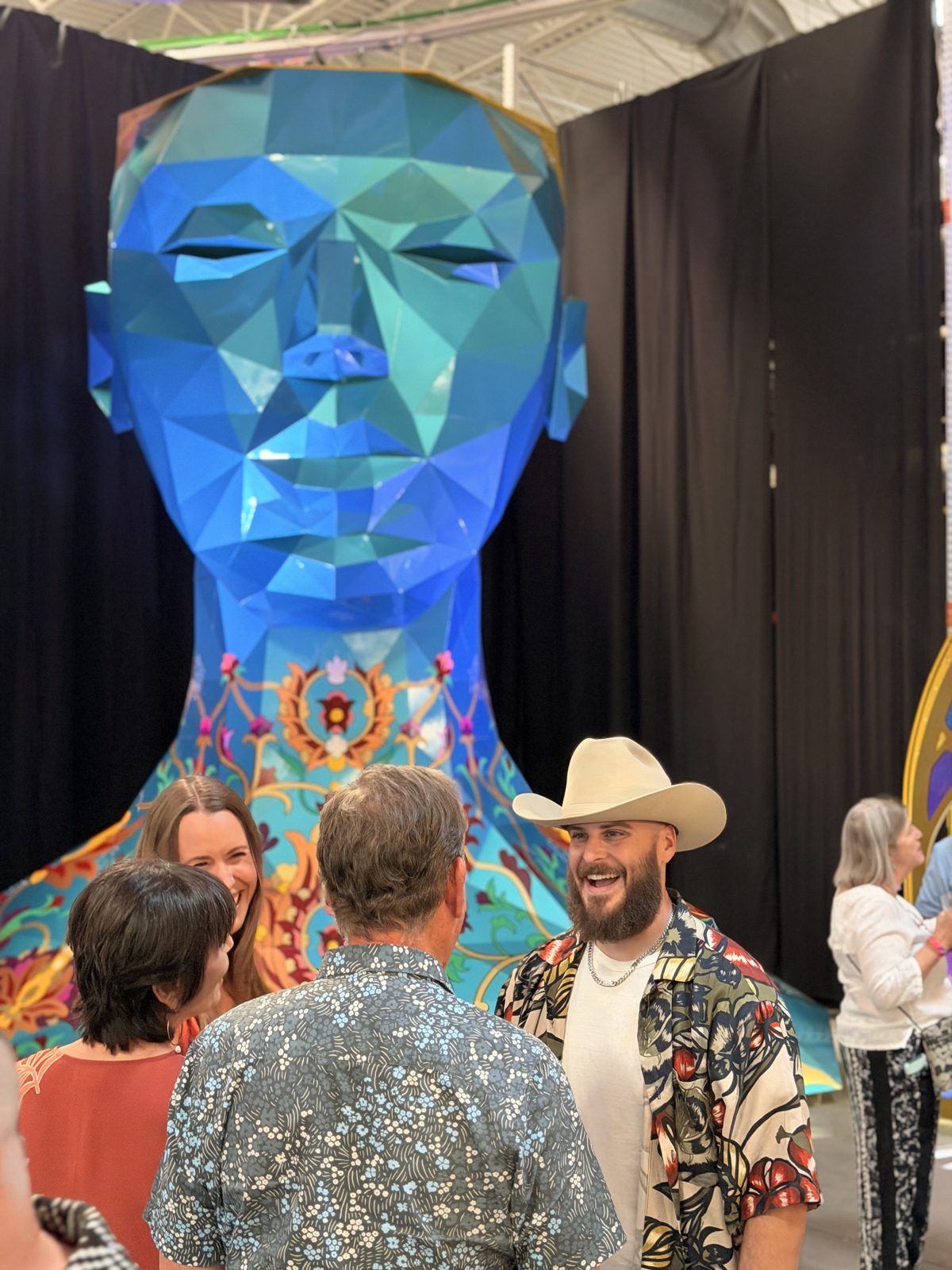 People laughing in front of ZARVAN, a 28 foot faceted blue head sculpture by Gazelle Dasti